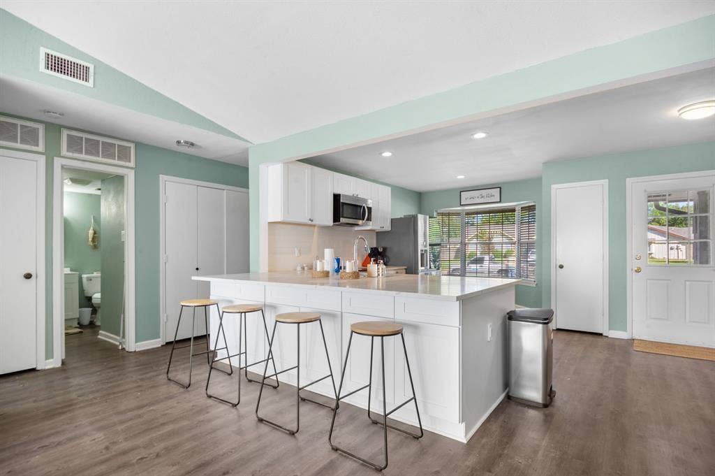 Irving, TX 75060 Kitchen featuring white cabinetry, a large kitchen island with counter seating, stainless steel appliances, wood-finish flooring, and a large window