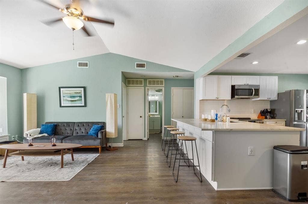 Irving, TX 75060 Vaulted ceilings with a ceiling fan, wood-finish flooring, white kitchen cabinetry, stainless steel appliances, and a kitchen island with seating