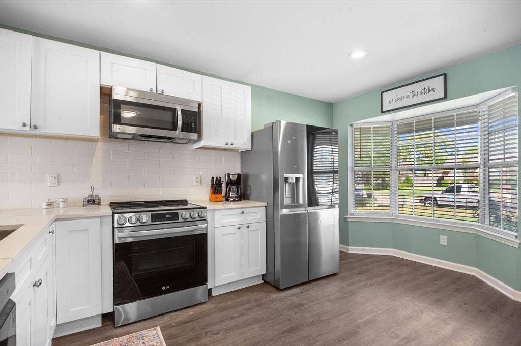 Irving, TX 75060 Modern kitchen featuring white cabinetry, stainless steel appliances, a subway tile backsplash, wood-finish flooring, and a bay window