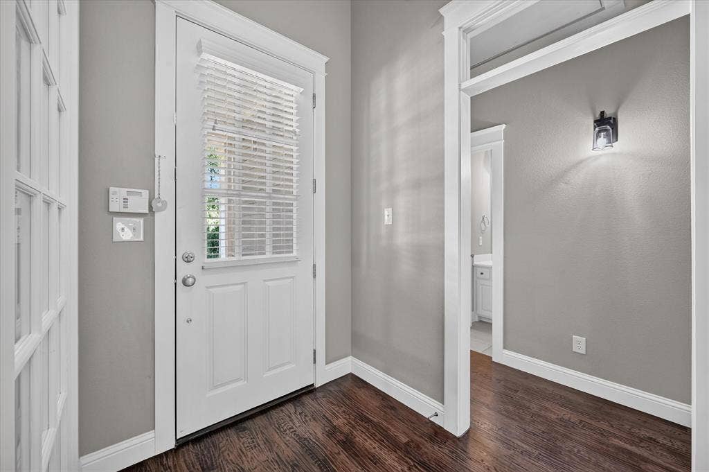 Rich wood-finish flooring with crisp white trim, a transom window door with privacy blinds, and a built-in wall sconce
