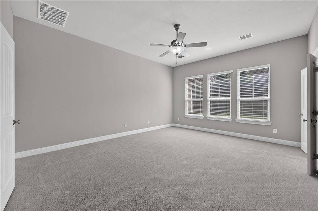 Spacious room featuring gray carpet, three large windows with blinds, a ceiling fan, and white trim