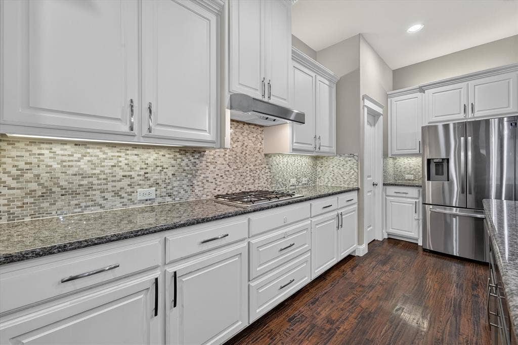 White cabinetry with chrome hardware, dark wood-finish flooring, stainless steel appliances, granite countertops, and mosaic tile backsplash