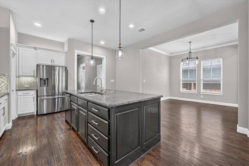 Sophisticated kitchen featuring a substantial center island with dark cabinetry and granite countertop, stainless steel appliances, white perimeter cabinetry, pendant lighting, and warm wood-finish flooring throughout the open-concept space