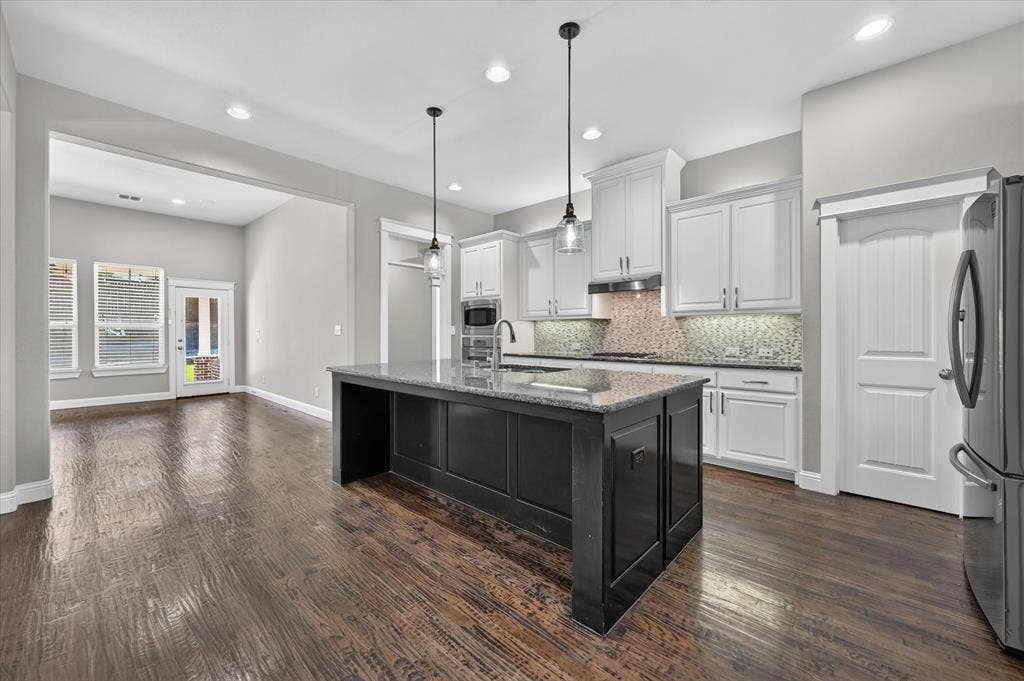 Open-concept kitchen featuring a large central island with a granite countertop, white cabinetry, integrated stainless steel appliances, and wood-finish flooring