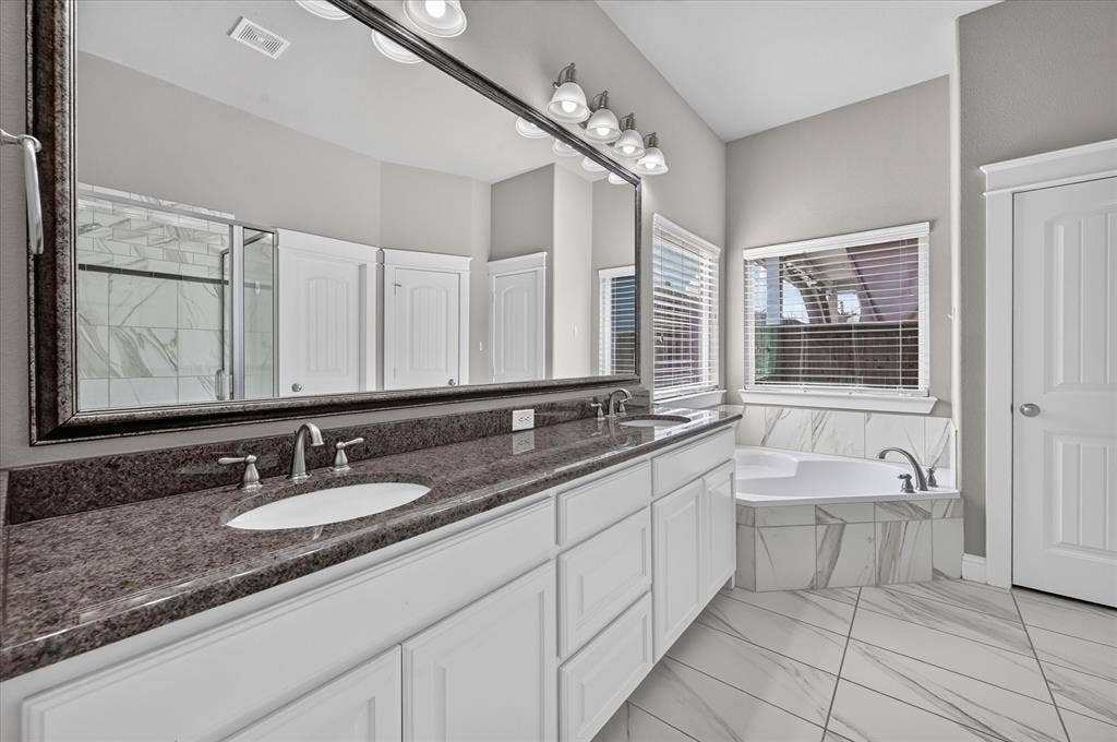 Dual vanity with undermount sinks and dark stone countertops, white cabinetry, large framed mirror, built-in tub with tile surround, and large format tile flooring