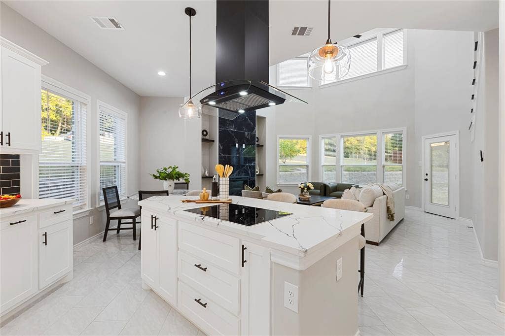 702 Linkcrest Drive, Duncanville, TX 75137 Open-concept living space with a kitchen island featuring an integrated cooktop, white cabinetry with dark hardware, a modern range hood, and high ceilings with upper windows