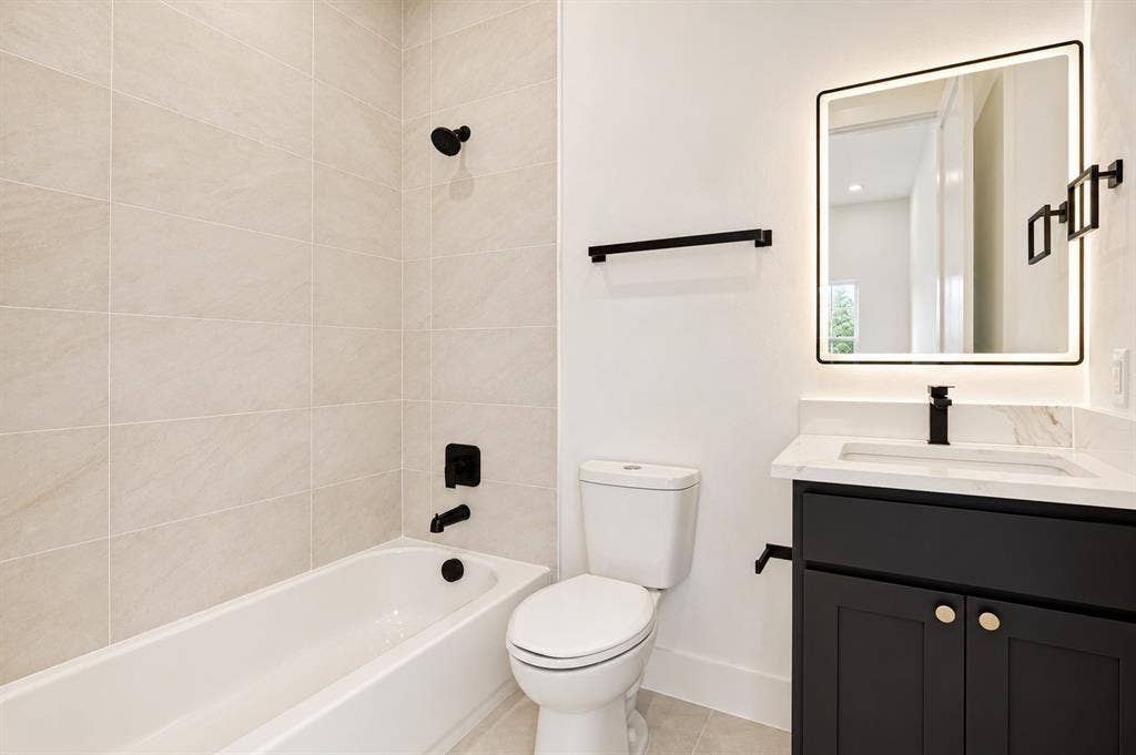 314 Avenue L, Dallas, TX 75203 Soaking tub with matte black fixtures, neutral-toned tile surround, single vanity with black cabinetry, white countertop, and illuminated framed mirror