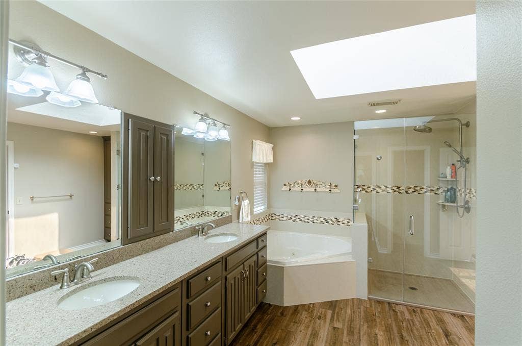 4009 Wilshire Court, Plano, TX 75023 Dual vanity with integrated sinks, a built-in jetted tub, glass-enclosed shower, wood-finish flooring, and a ceiling-mounted skylight