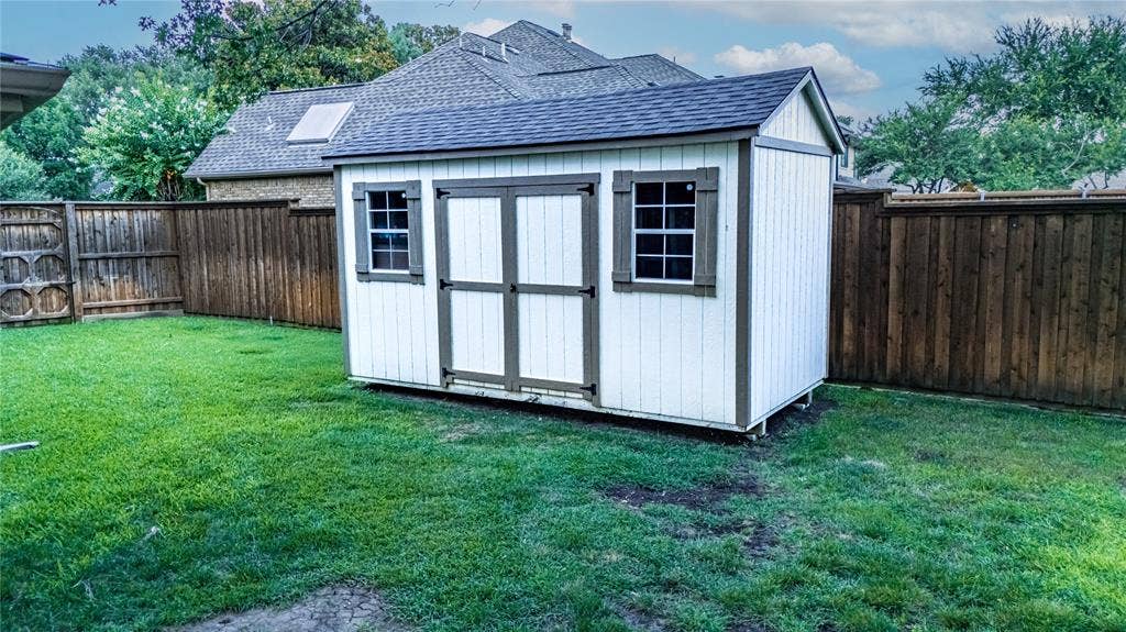 4009 Wilshire Court, Plano, TX 75023 White exterior shed with a dark shingle roof, double entry doors, two windows with dark shutters, and a surrounding wood privacy fence