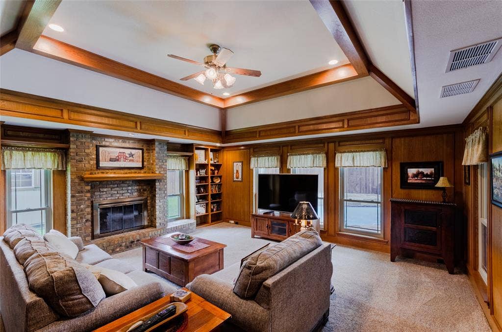 4009 Wilshire Court, Plano, TX 75023 Spacious living area featuring a brick fireplace, built-in shelving, wood-paneled walls, a tray ceiling with wood beams, and a ceiling fan with integrated lighting