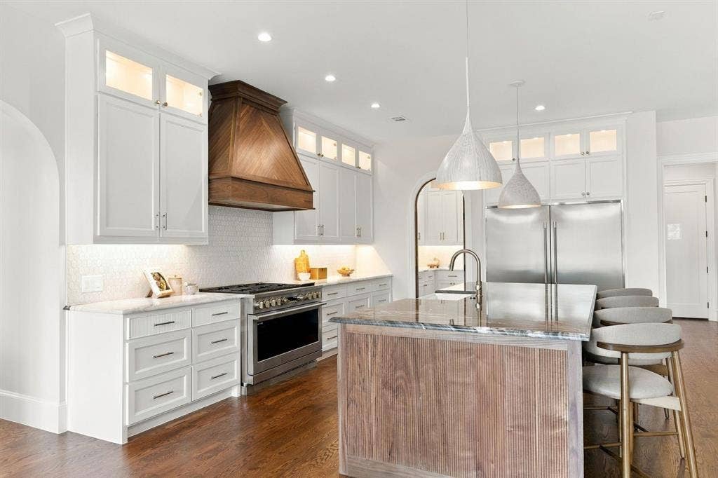 3368 Dorado Beach Drive, Farmers Branch, TX 75234 Gourmet kitchen featuring a wood-panel range hood, stainless steel appliances, white shaker cabinetry, and a large center island with a wood-finish base and stone countertop
