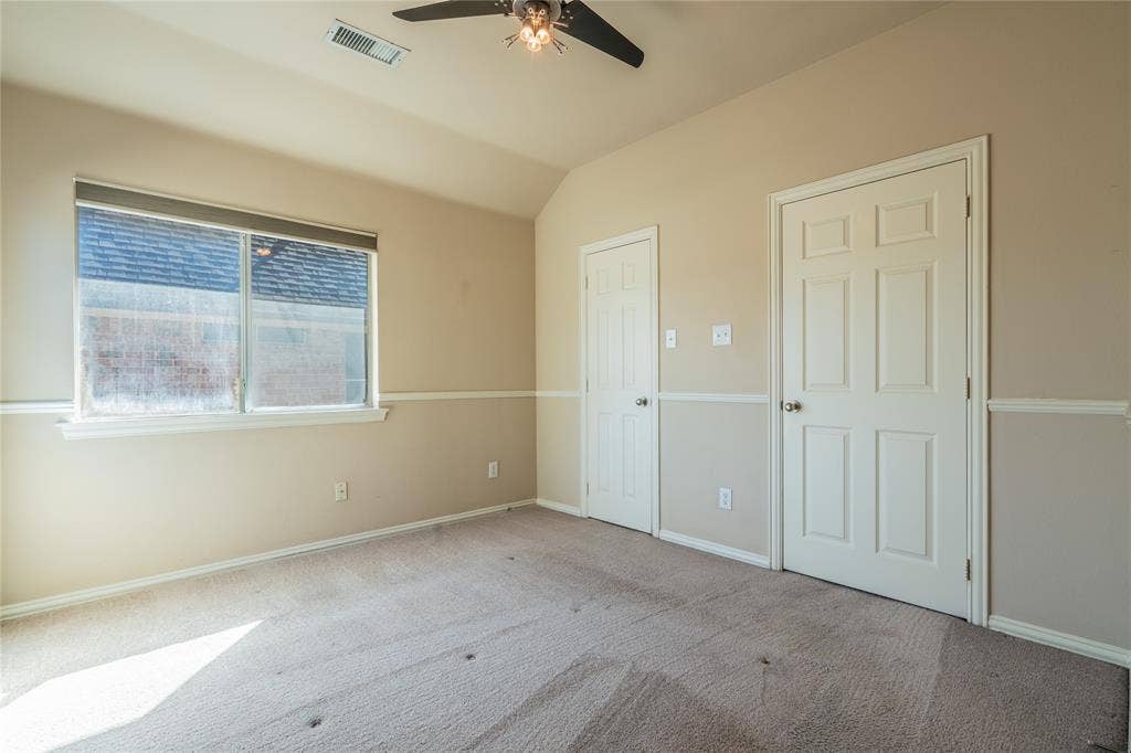 Another view of the bedroom with a vaulted ceiling with a ceiling fan, window with roller shades, two panel doors, and neutral carpeting