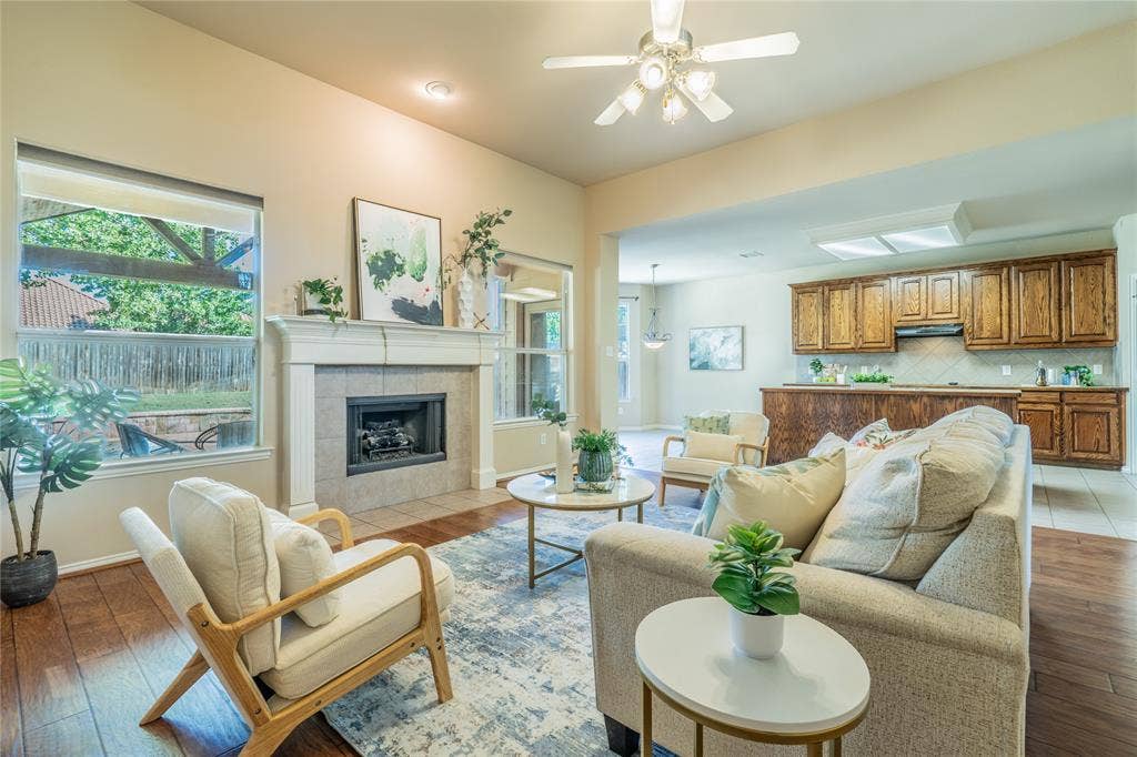 Living area featuring wood-finish flooring, a fireplace with a tiled surround and mantle, a ceiling fan, and large windows