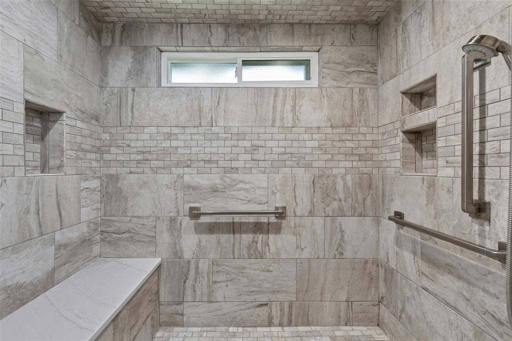 Oversized Walk-In Shower — The remodeled primary shower features expansive tilework, built-in niches, a bench, handheld shower fixture, and grab bars for added convenience and flexibility.