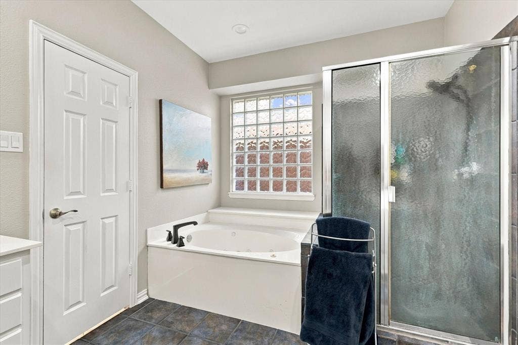 Soaking tub with matte black fixtures, glass block window, glass-enclosed shower, and dark stone-finish floor tile