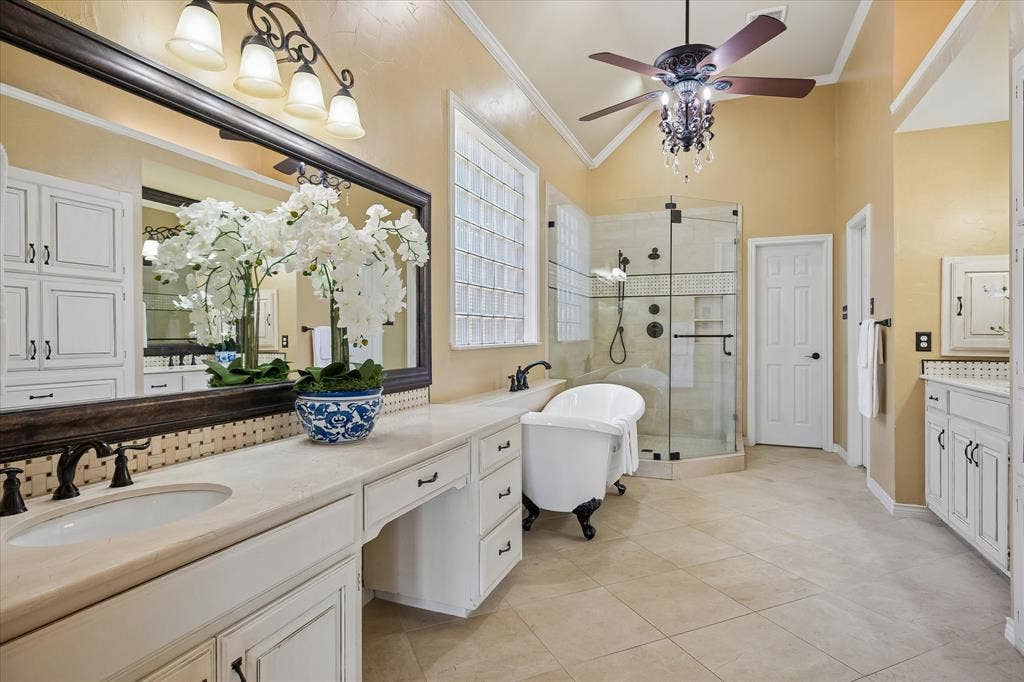 2204 Creekside Circle S, Irving, TX 75063 Spa-inspired primary bathroom featuring dual vanities with white cabinetry and marble countertops, bronze fixtures, a freestanding soaking tub, walk-in shower with frameless glass enclosure and tile surround, ceiling fan with integrated lighting, and neutral porcelain tile flooring