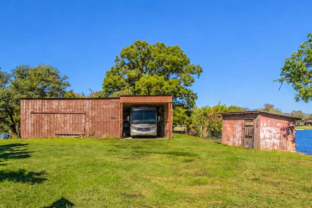 118 Spring Lake Drive, Lacy Lakeview, TX 76705 Expansive grassy property featuring a large double level metal workshop with electricity and a RV storage area without electricity, a well shed, and mature trees. The shoreline extends behind the workshop on both sides.