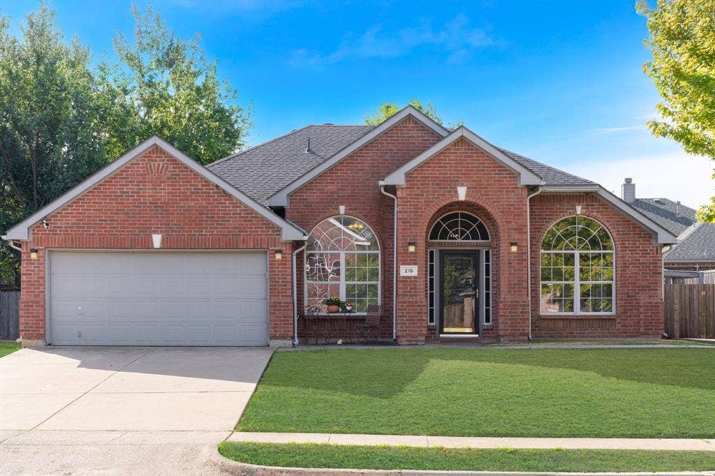 215 Longhorn Trail, Grand Prairie, TX 75052 Red brick exterior featuring a dark composition shingle roof, arched transom windows, and an attached two-car garage