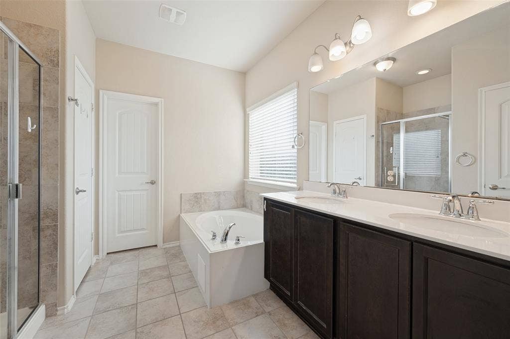 9801 Moccasin Creek Lane, McKinney, TX 75071 Dual vanity with dark wood-finish cabinetry and white countertops, a built-in soaking tub, a separate glass-enclosed shower, and neutral tile flooring