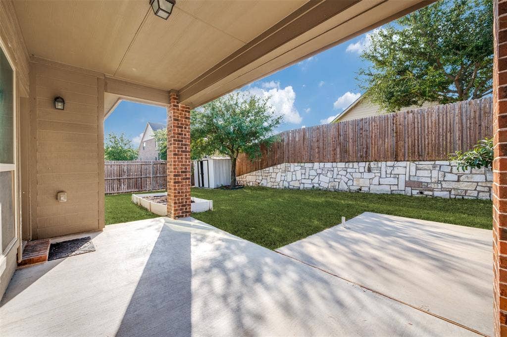 9801 Moccasin Creek Lane, McKinney, TX 75071 Covered patio with a concrete surface, brick columns, and a landscaped backyard featuring a stone retaining wall and a wooden privacy fence