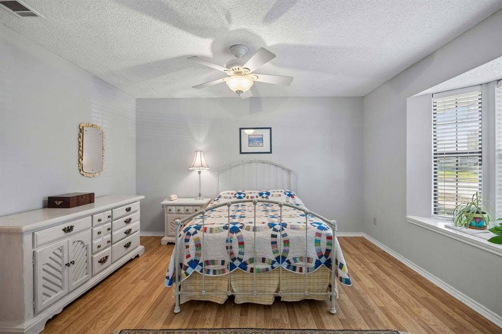1902 Wyatt Drive, Garland, TX 75040 Bright secondary bedroom featuring hardwood flooring, ceiling fan with integrated lighting, white six-drawer dresser with decorative hardware, and large window with plantation shutters providing natural light and privacy