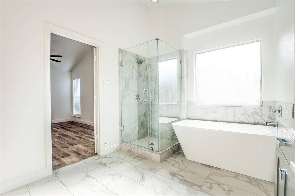 8341 Edgewater Drive, Frisco, TX 75034 Spacious bathroom featuring a frameless glass shower, freestanding soaking tub, large window, and marble-look tile flooring