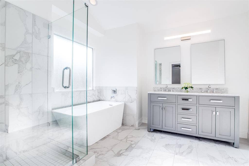 8341 Edgewater Drive, Frisco, TX 75034 Freestanding soaking tub, glass-enclosed shower, dual vanity with gray cabinetry, wall-mounted light fixture, and marble-patterned tile flooring