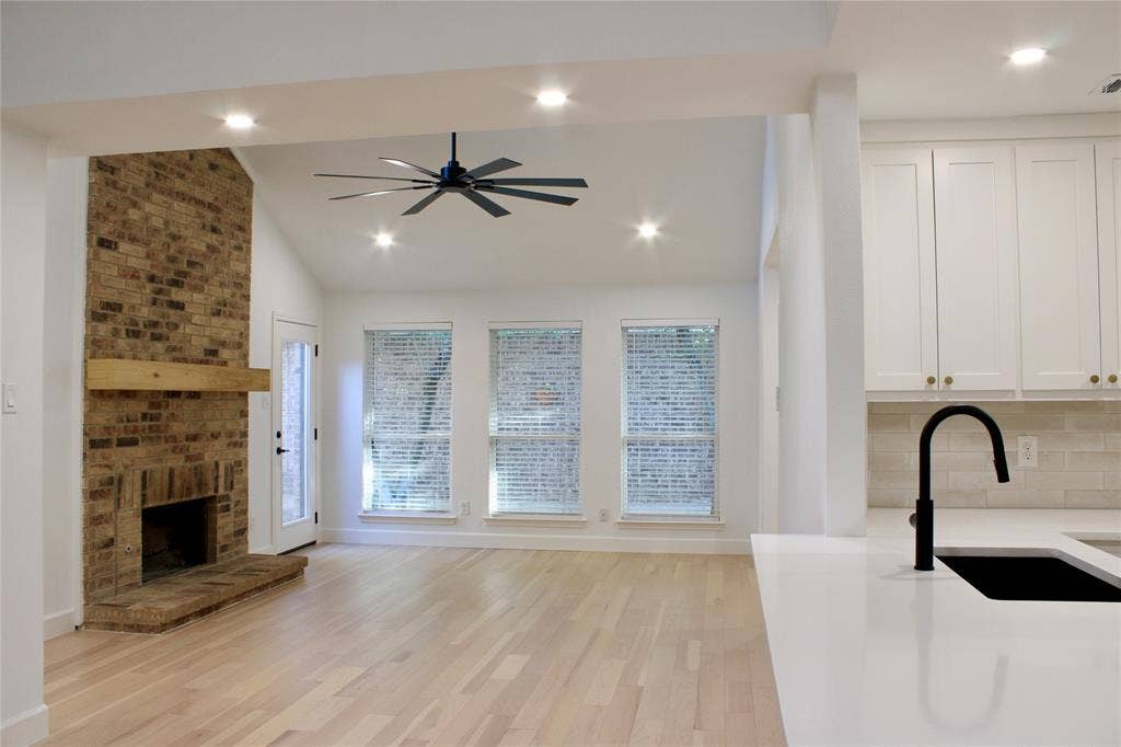3706 Aries Drive, Garland, TX 75044 Vaulted ceiling with a ceiling fan, brick fireplace with a wood mantle, light wood-finish flooring, three windows, and white kitchen cabinetry with a matte black faucet