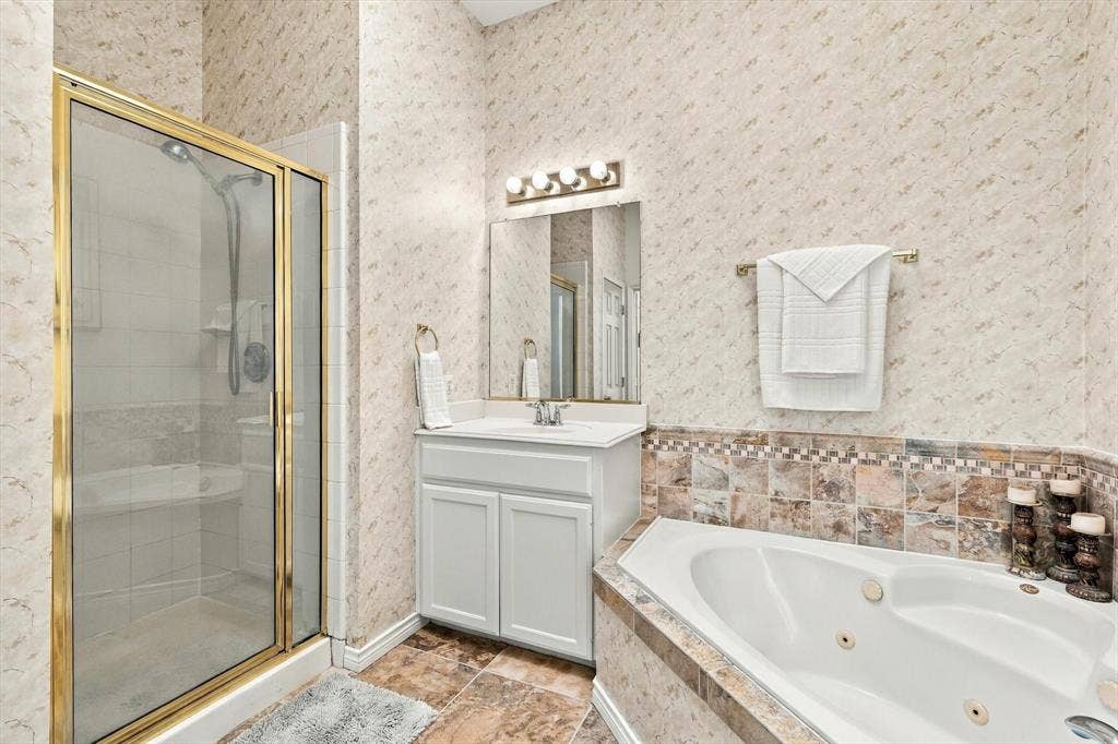 Shower with gold-toned frame and clear glass enclosure, single vanity with white cabinetry, built-in mirror with integrated lighting, tiled flooring, and corner jetted bathtub with tiled surround