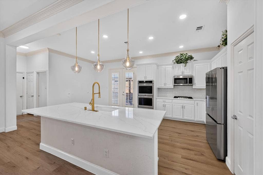 10601 Whispering Pines Drive, Frisco, TX 75033 Kitchen featuring a large island with a surface-mount sink and gold-finish faucet, white cabinetry, integrated stainless steel appliances, and wood-finish flooring