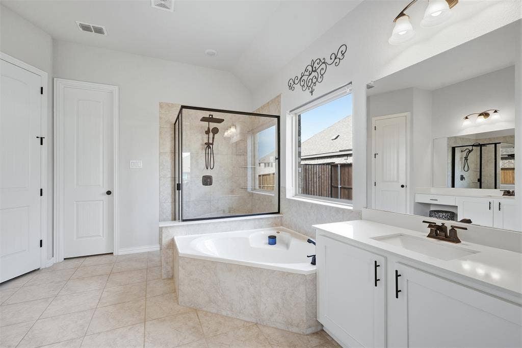 Corner soaking tub, glass-enclosed shower with dark fixtures, white vanity with integrated sink, rectangular window, and light-toned tile flooring