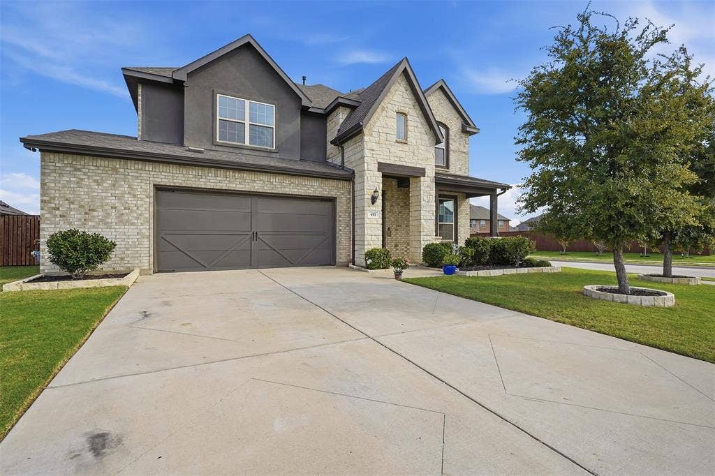 Stone and brick exterior with stucco accents, dual garage doors, covered entry, and landscaped front yard with mature trees