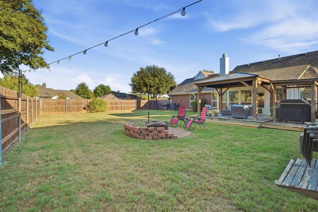 506 Amy Jo Circle, Aubrey, TX 76227 Expansive backyard featuring a manicured lawn, full perimeter fencing, a covered patio structure, and a brick-lined fire pit