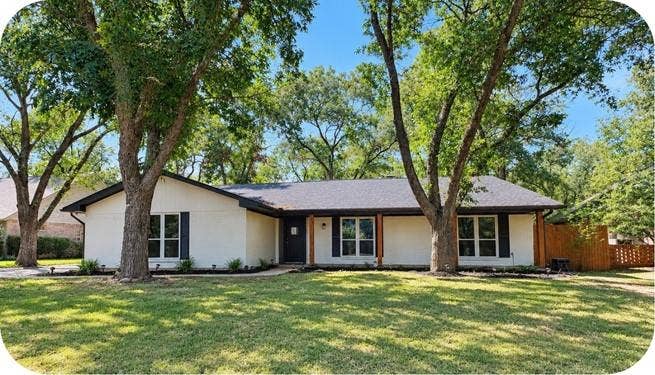 8513 Ravenswood Road, Granbury, TX 76049 White painted brick exterior with black window shutters, dark shingled roof, mature trees, and a wooden privacy fence