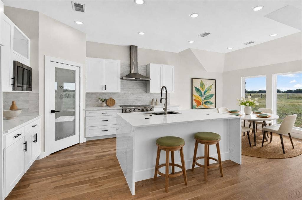 15166 FM 3133, Anna, TX 75409 Modern kitchen featuring white cabinetry, a large center island with a sink, stainless steel range hood, wood-finish flooring, and a dining area with multiple windows - Virtually Staged