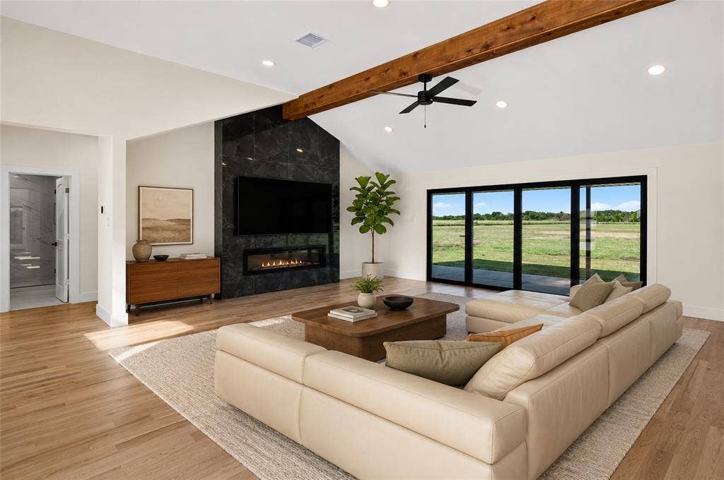 15166 FM 3133, Anna, TX 75409 Spacious living area featuring a vaulted ceiling with an exposed wood beam, a black marble-style fireplace, and wide plank wood-finish flooring - Virtual Staging
