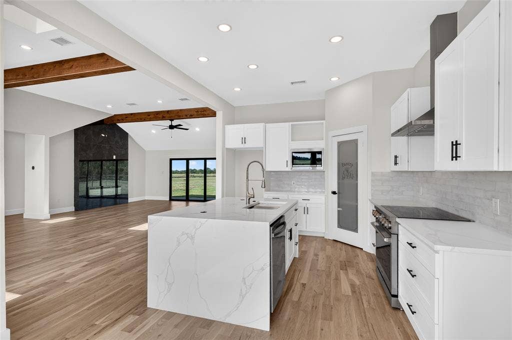 15166 FM 3133, Anna, TX 75409 Open-concept kitchen featuring a waterfall island with an integrated sink, white cabinetry with matte black hardware, stainless steel appliances, and wood-finish flooring