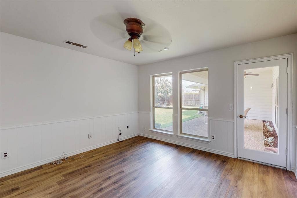 Wood-finish flooring, white panel wainscoting, ceiling fan with light fixture, large windows, and a glass-paneled door leading to an outdoor space