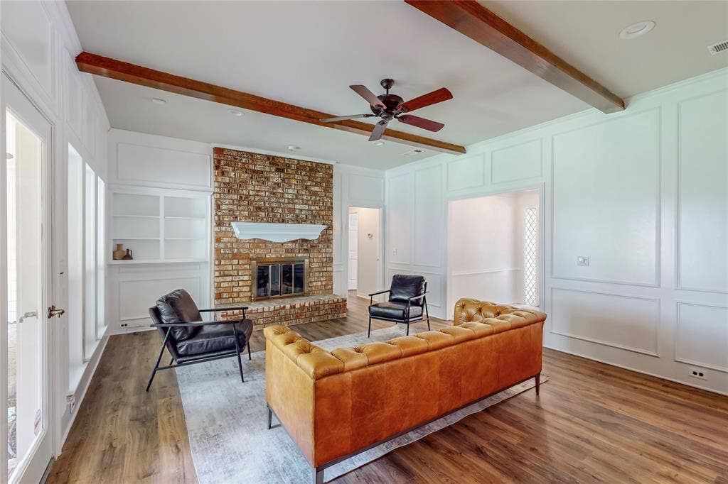 Brick fireplace with a classic mantel, wood-finish flooring, white built-in shelving, decorative wall paneling, and exposed ceiling beams
