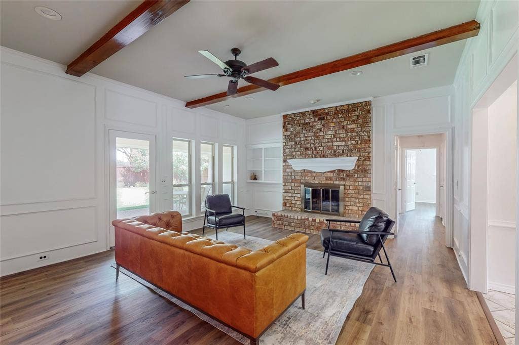 Living area featuring exposed ceiling beams, wood-finish flooring, brick-faced fireplace with hearth, built-in shelving, and multiple windows