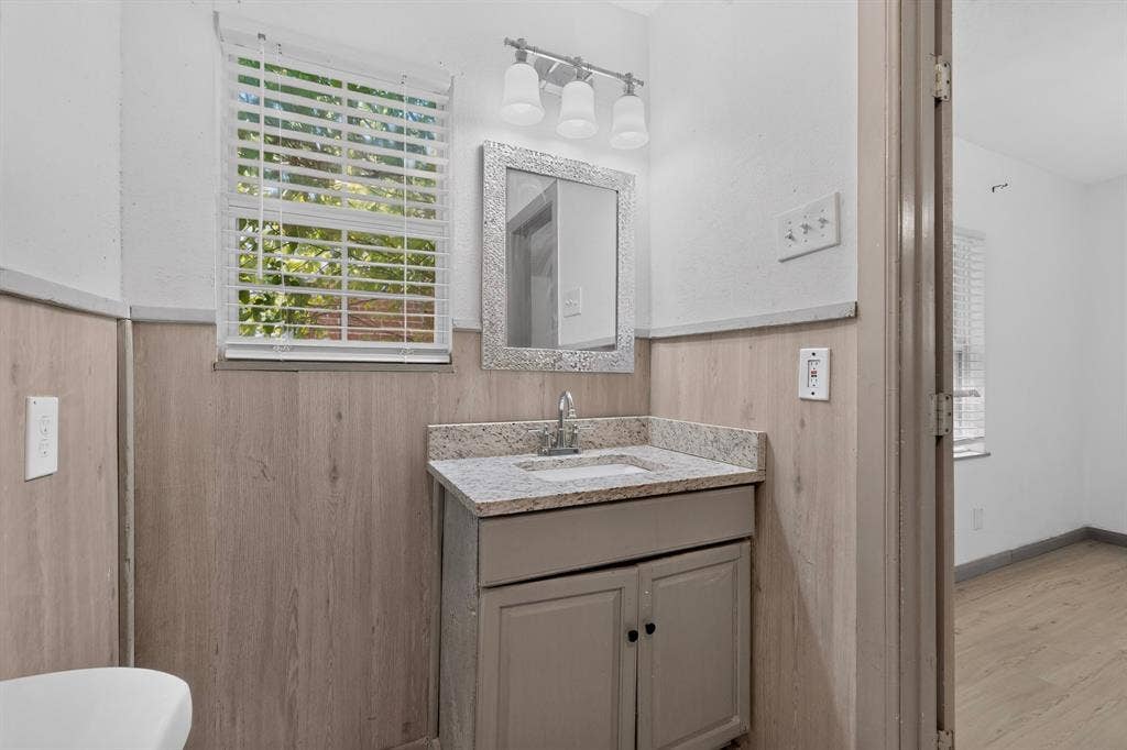 Vanity featuring stone-style countertop with integrated basin, light-toned cabinetry, three-light fixture, and wood-finish wall paneling