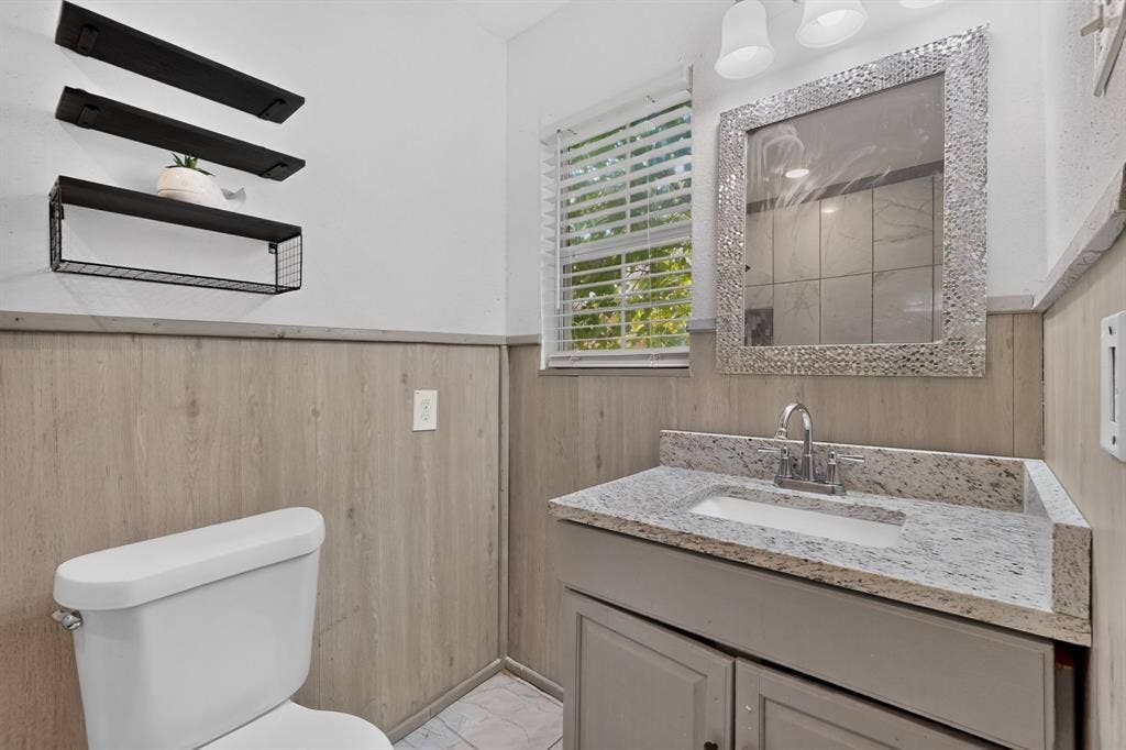 Bathroom vanity featuring a stone-composite countertop, undermount sink with a metallic faucet, wood-finish wainscoting, and three wall-mounted metal shelves
