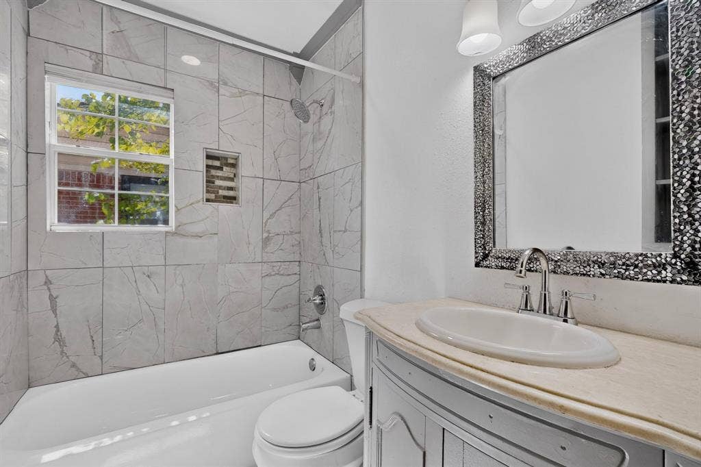 Bathroom featuring a tub-shower combination with floor-to-ceiling large-format tile surround, recessed decorative niche, neutral-toned countertop with an integrated oval basin, and wall-mounted vanity lighting