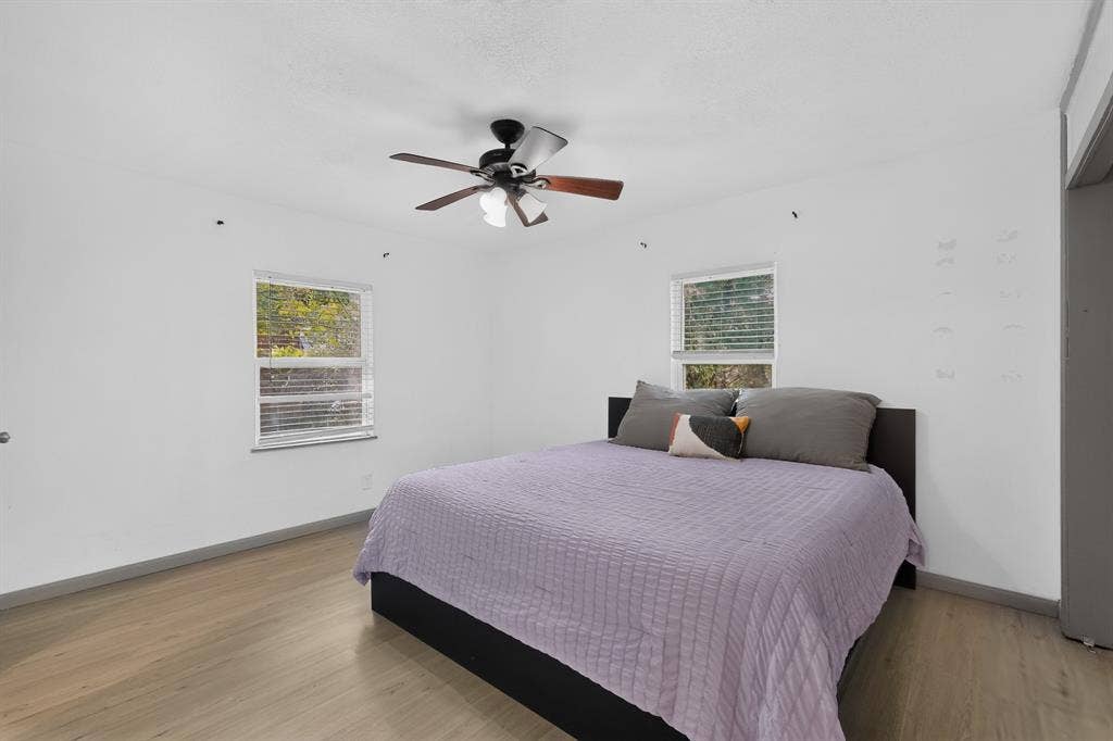 Neutral-toned interior space featuring light wood-finish flooring, two windows with horizontal blinds, and a central ceiling fan with integrated lighting