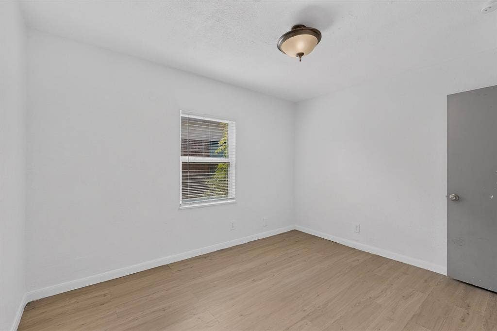 Interior space featuring wood-finish flooring, white walls, a single-window with horizontal blinds, and a neutral-toned door with a metallic knob