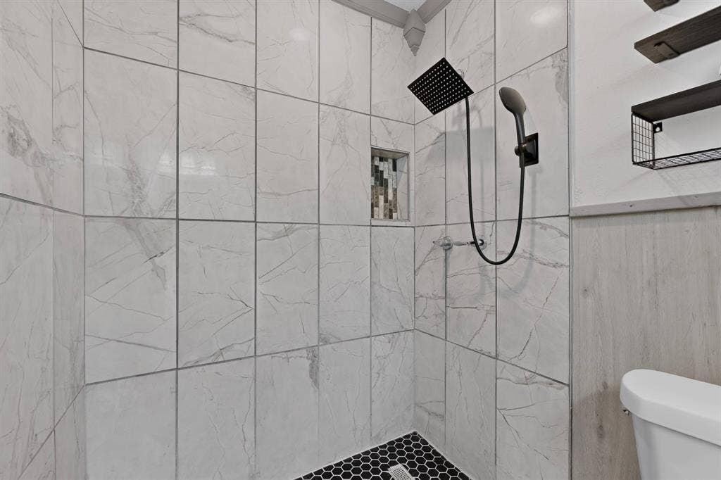 Walk-in shower featuring large-format stone-look tile walls, matte black rainfall showerhead with handheld attachment, built-in mosaic tile niche, and black hexagonal floor tile