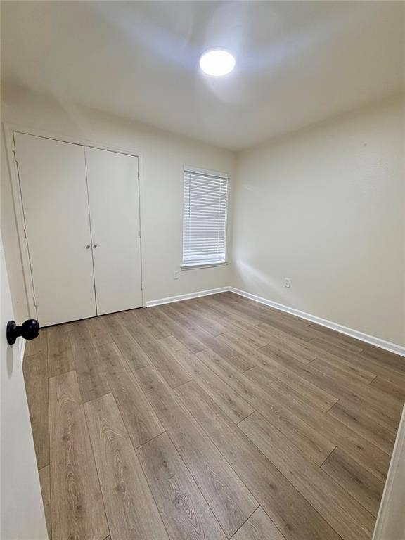 Wood-finish flooring, built-in closet with bifold doors, window with horizontal blinds, and a flush-mount ceiling light fixture