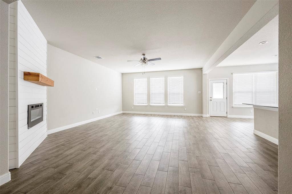 1548 Seabiscuit Drive, Granbury, TX 76049 Spacious living area featuring wood-finish flooring, a fireplace with a wood mantle, multiple windows, a ceiling fan, and a light-colored entry door