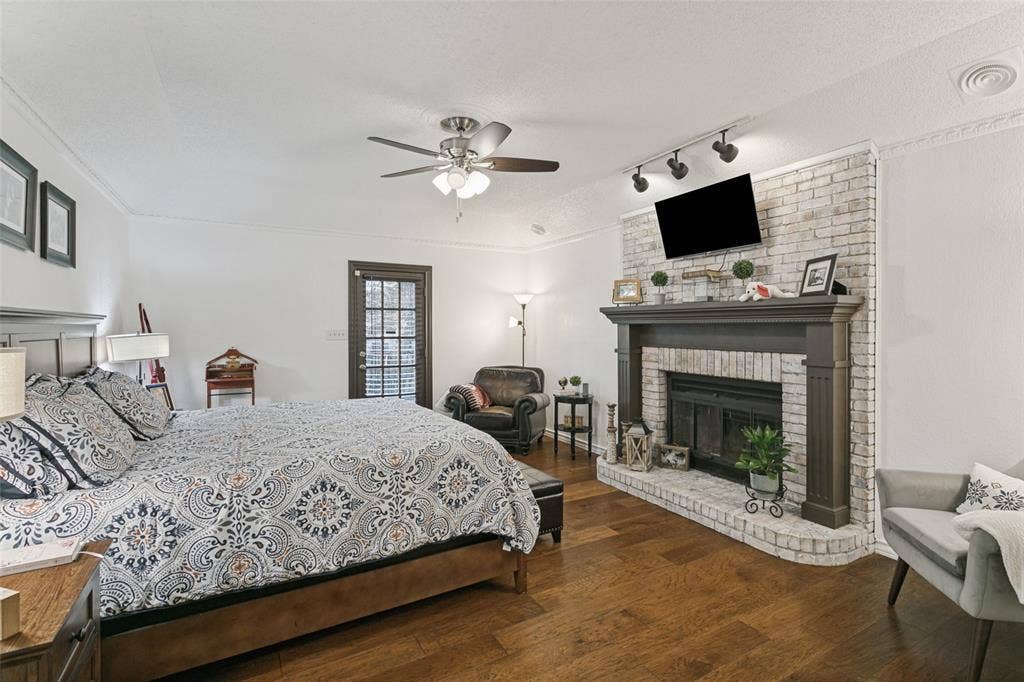 6801 Bellaire Court S, Benbrook, TX 76132 Primary bedroom with Rich wood-finish flooring, a white brick fireplace with a dark wood mantel, a ceiling fan, crown molding, and a paneled glass door