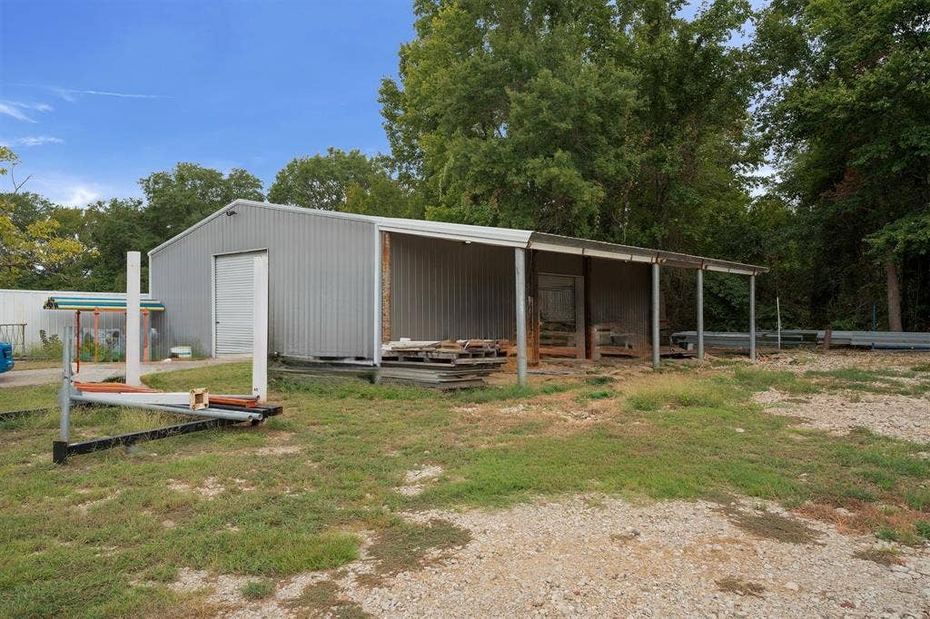 5989 State Highway 31 E, Murchison, TX 75778 Metal-sided building featuring a large roll-up door, an attached open-sided covered area, and a gravel surface